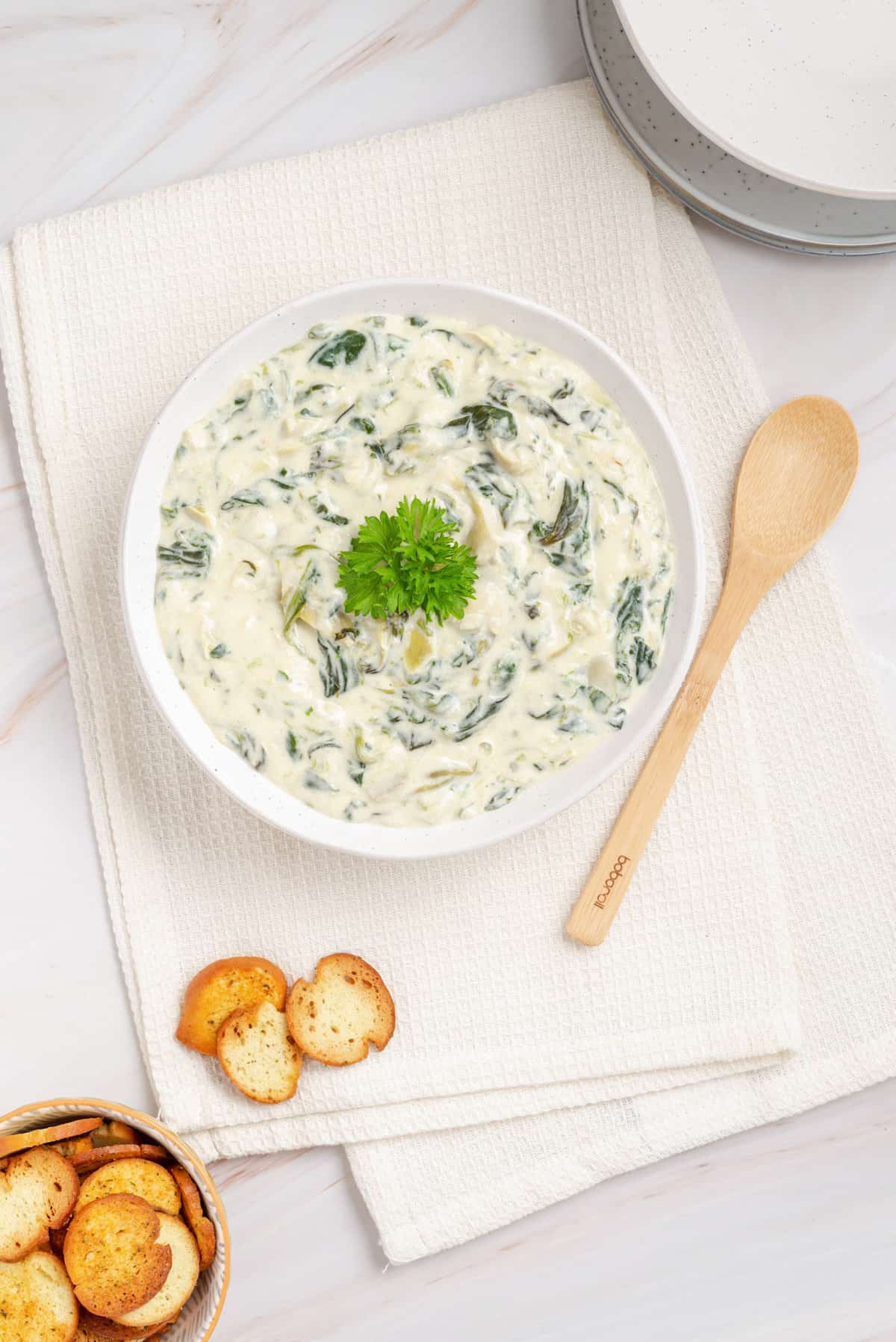 Overhead view of bowl of Instant Pot spinach artichoke dip with parsley garnish, crostini and wooden spatula on the side.