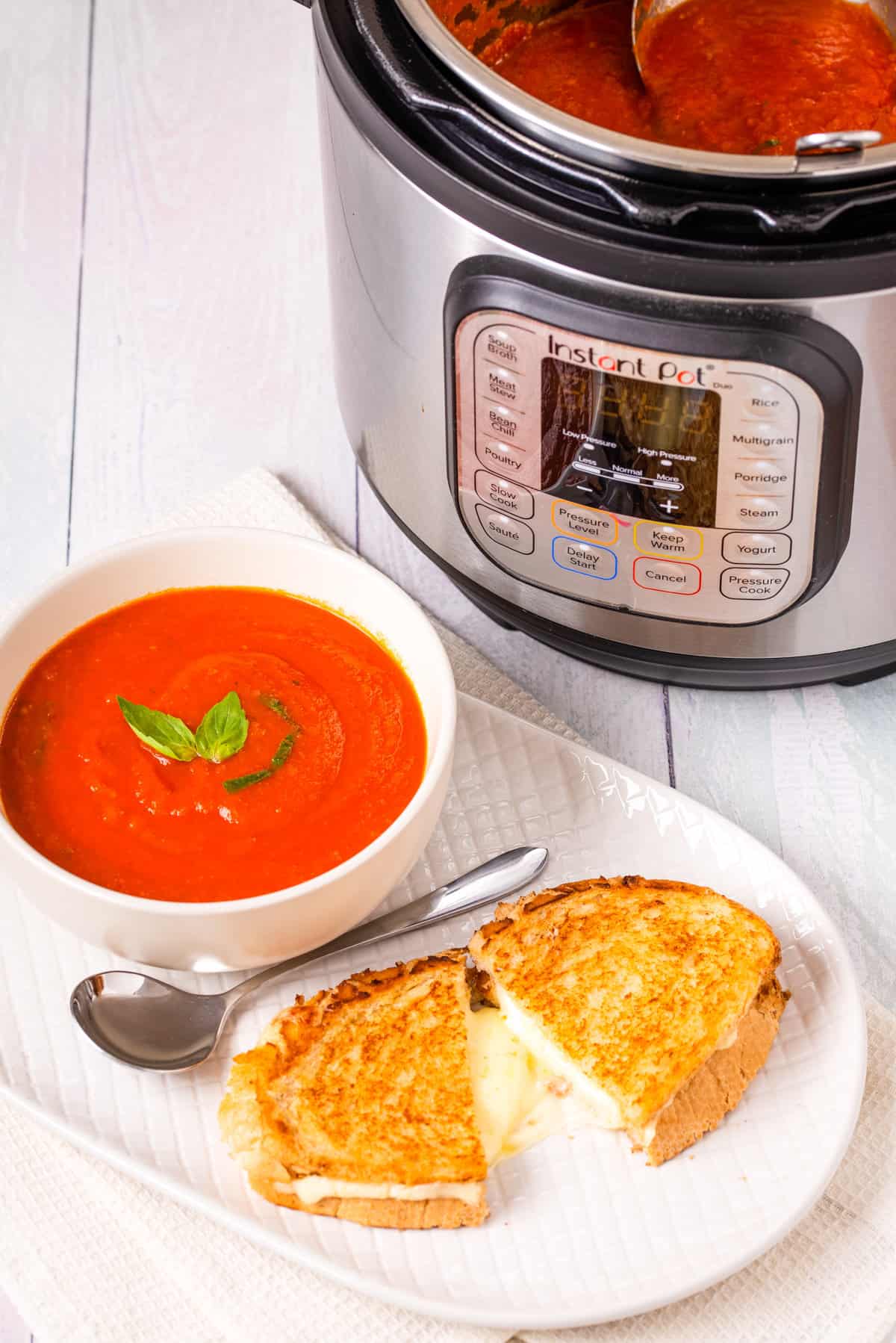 Close up shot of finished tomato soup in a white bowl, with a grilled cheese toastie on a white plate, instant pot in the background.