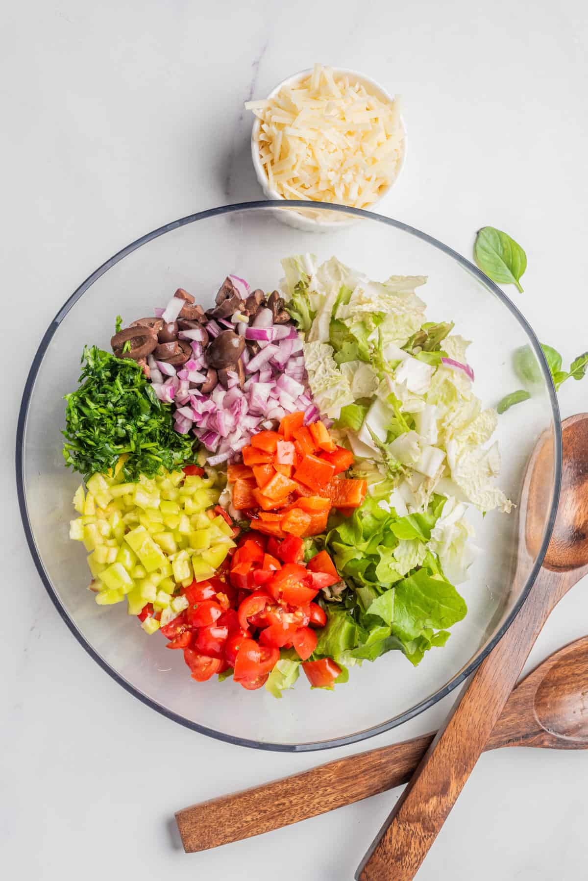 An image of Romaine lettuce, endive, red onion, cherry tomatoes, cucumber, pepperoncini, roasted red peppers, olives and parsley in a large mixing bowl. 