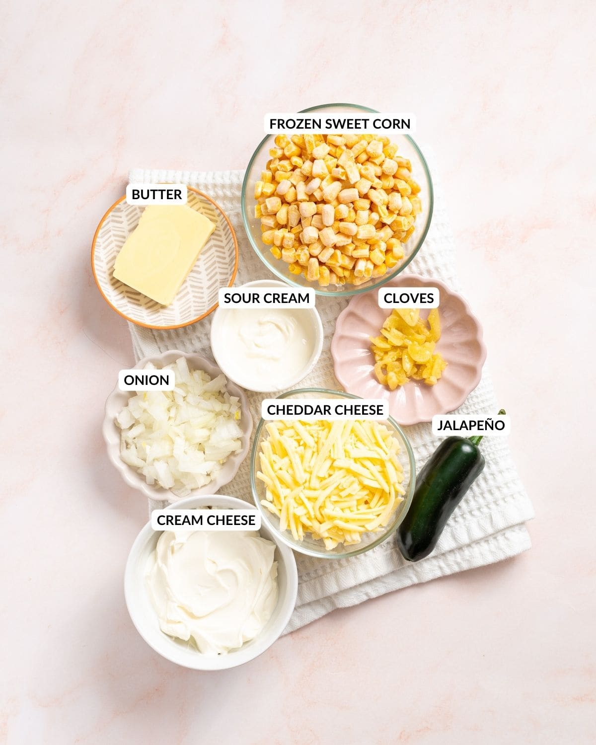 An image of the ingredients of jalapeno corn: sweet corn, jalapenos, sour cream, cream cheese, cheddar cheese, butter, garlic cloves, and onions in separate containers with labels.