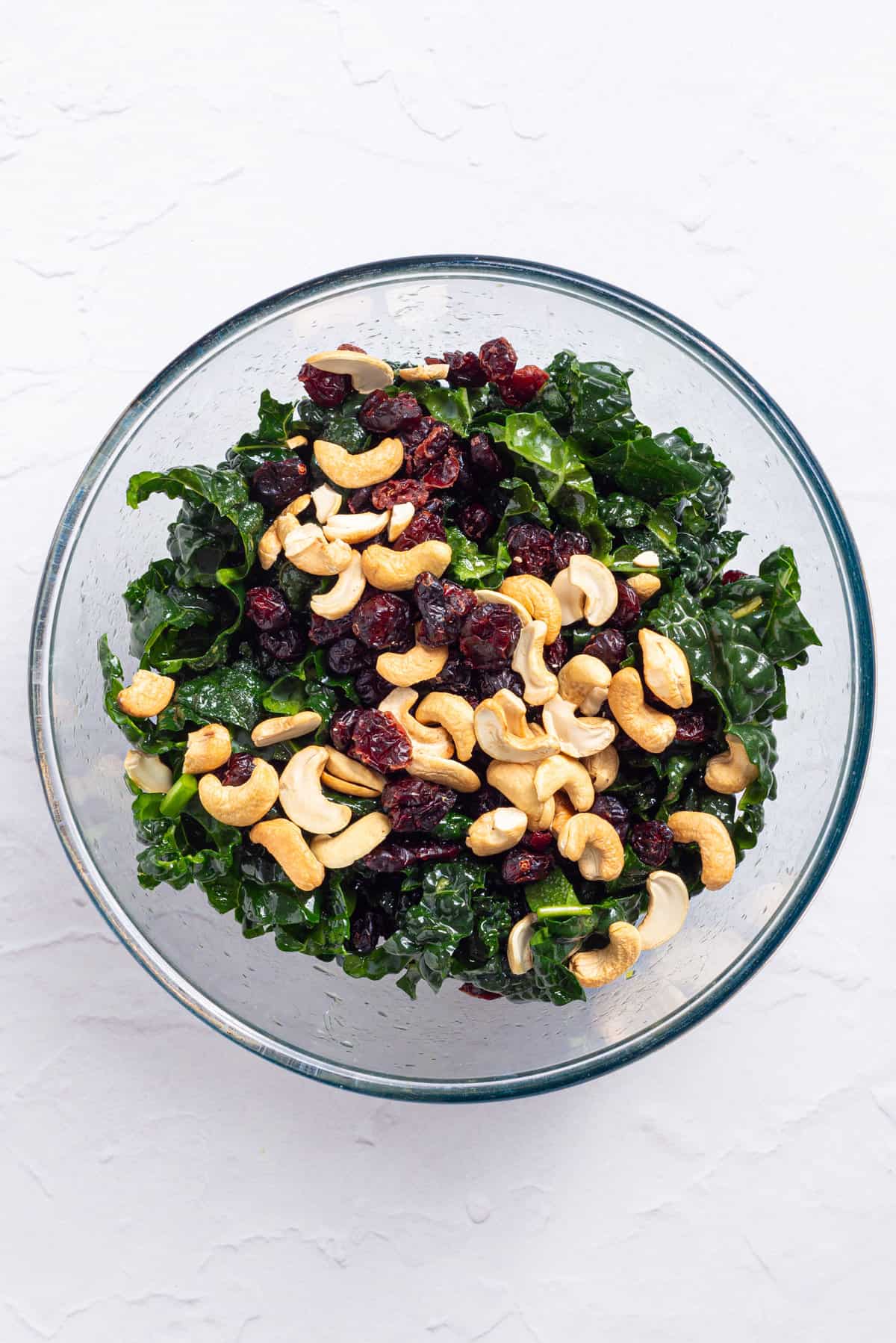 An image of kale leaves, cashews, and dried cranberries with salad dressing tossed in a bowl