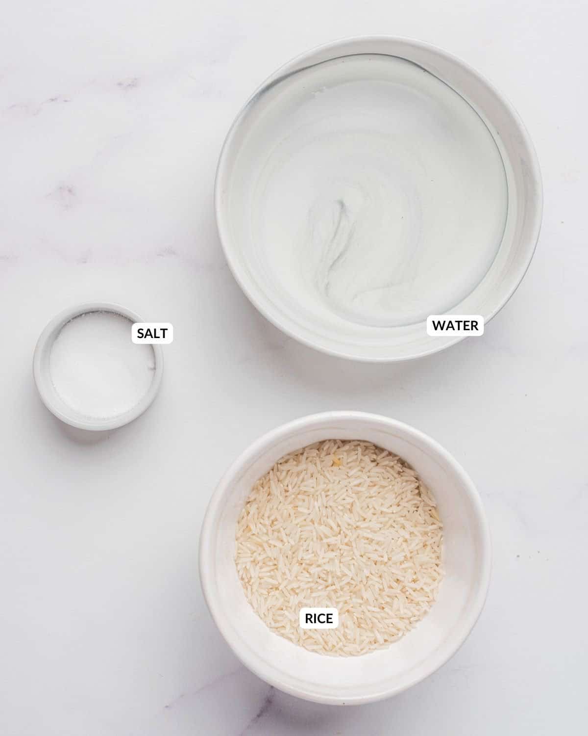 An overhead image of uncooked rice in one bowl, water in another bowl, and salt in a small bowl.
