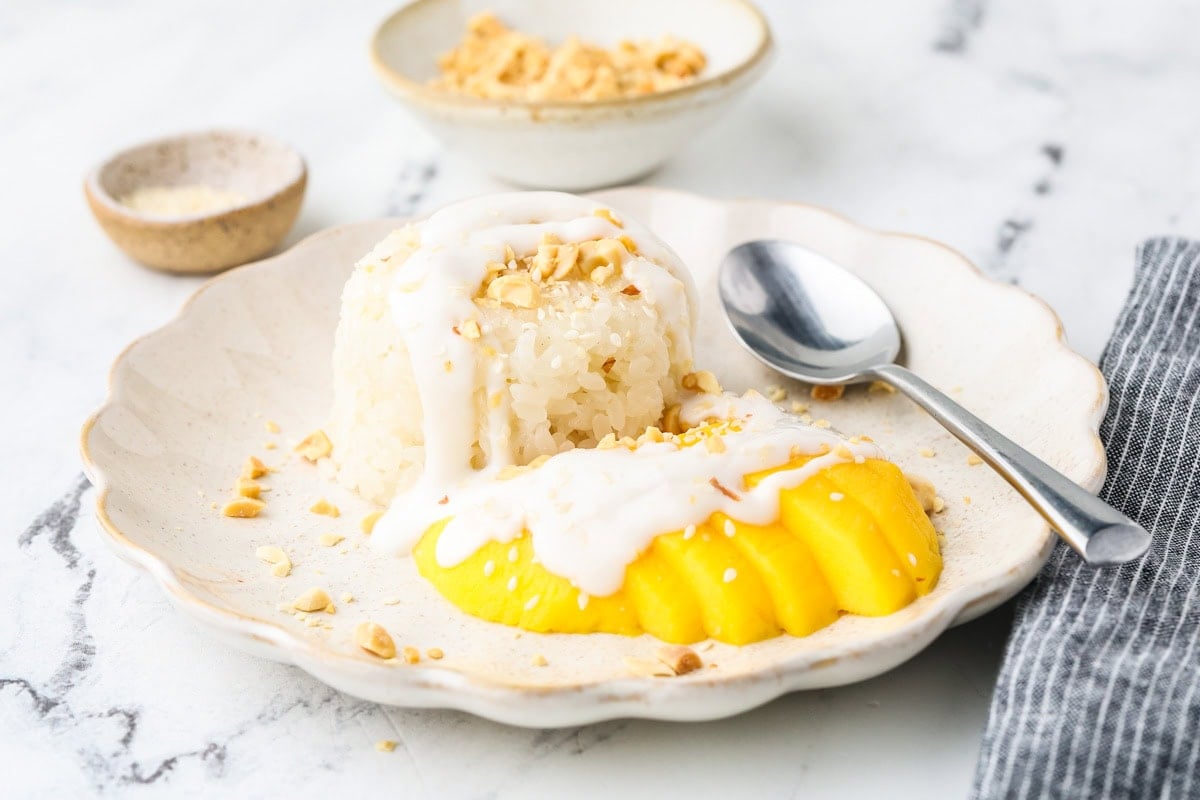 Mango sticky rice plated with coconut cream and mango slices, served with a spoon on a light marble background.