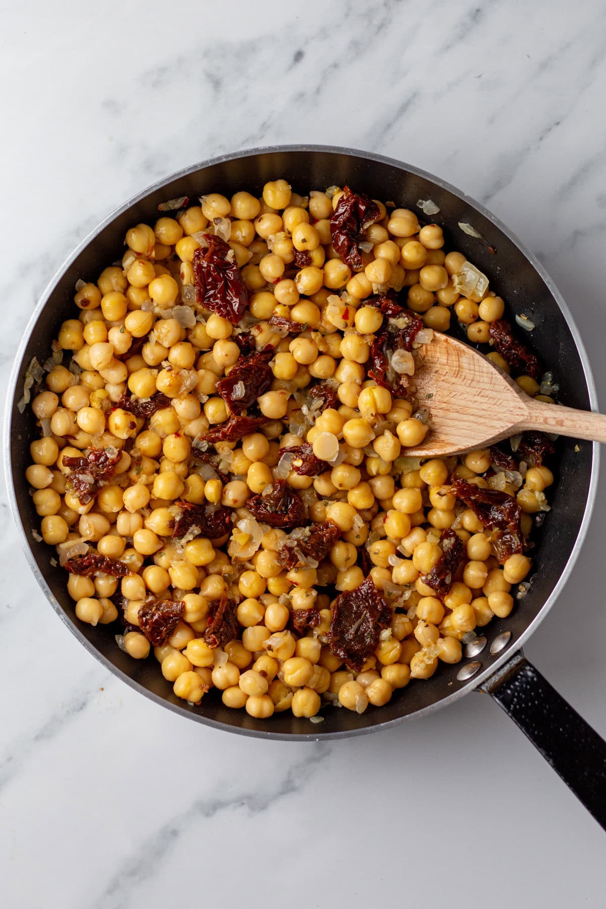 An overhead image of adding chickpeas and sun-dried tomatoes on a skillet.