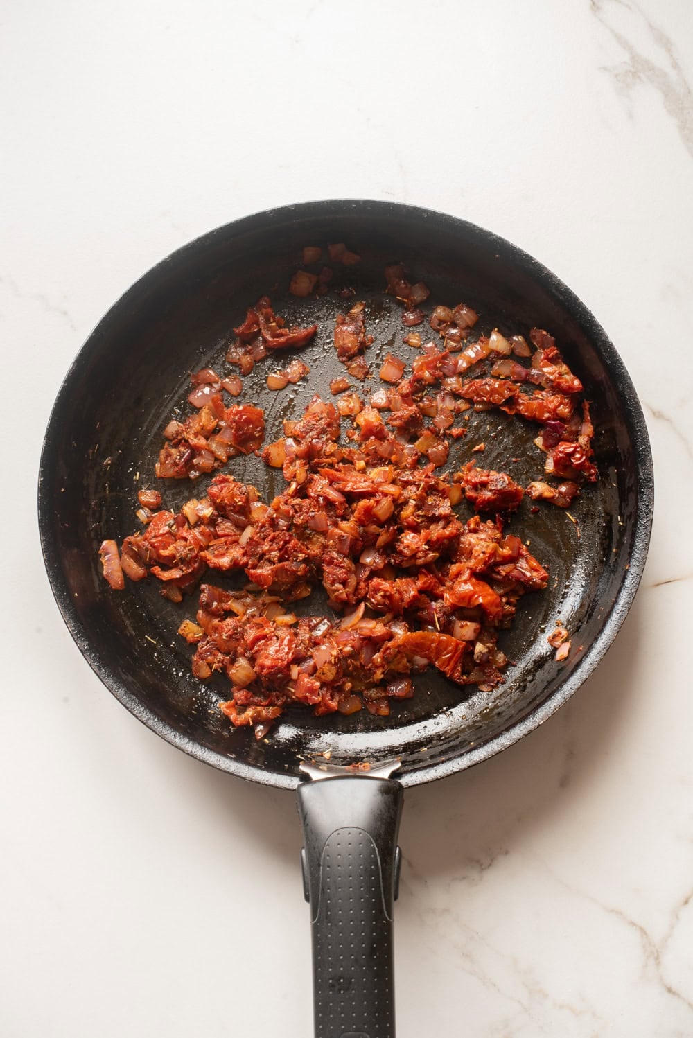 Diced onions, minced garlic, herbs, and darkened tomato paste sautéing together in a black skillet on a light marble surface.