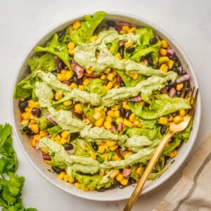 An overhead image of mexican corn salad with avocado lime dressing drizzled on top, and a fork resting on the side.