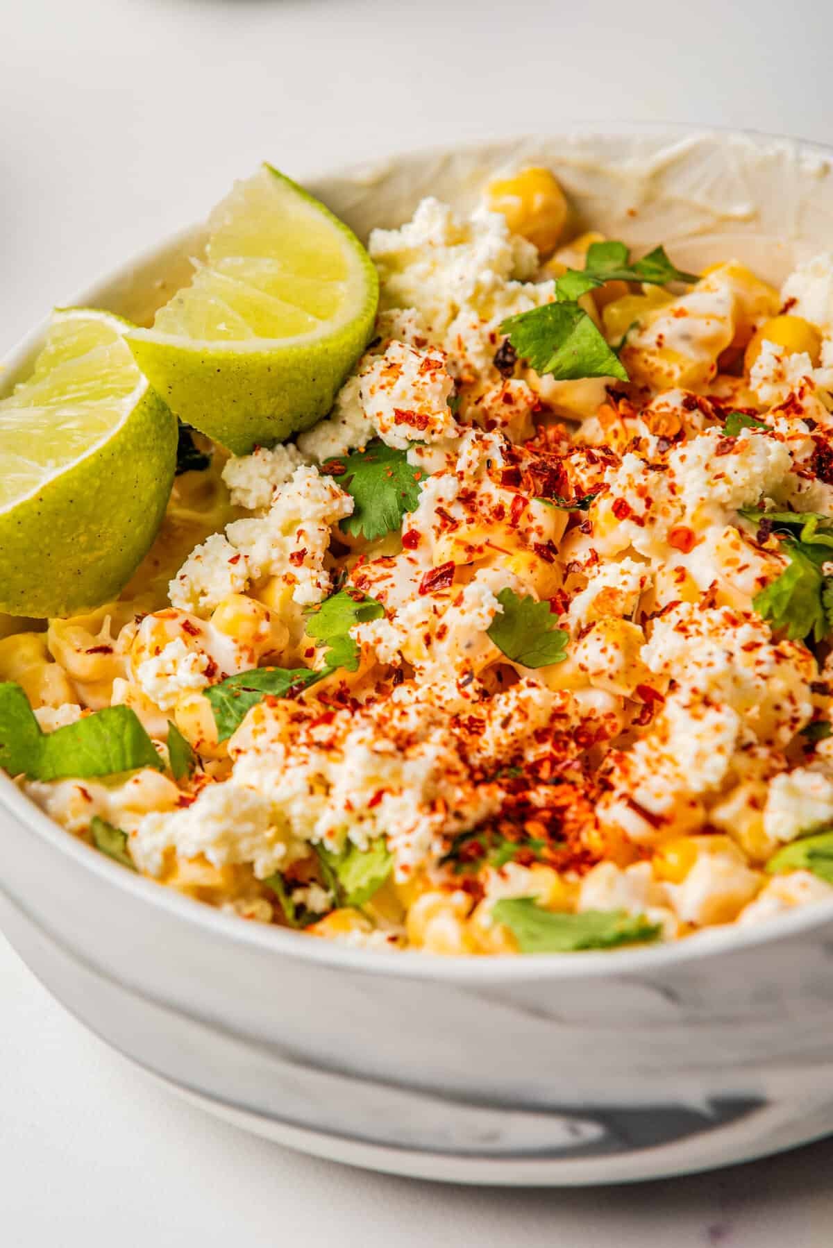 A close up image of Mexican street corn salad in a bowl with two slices of lime resting on the side.