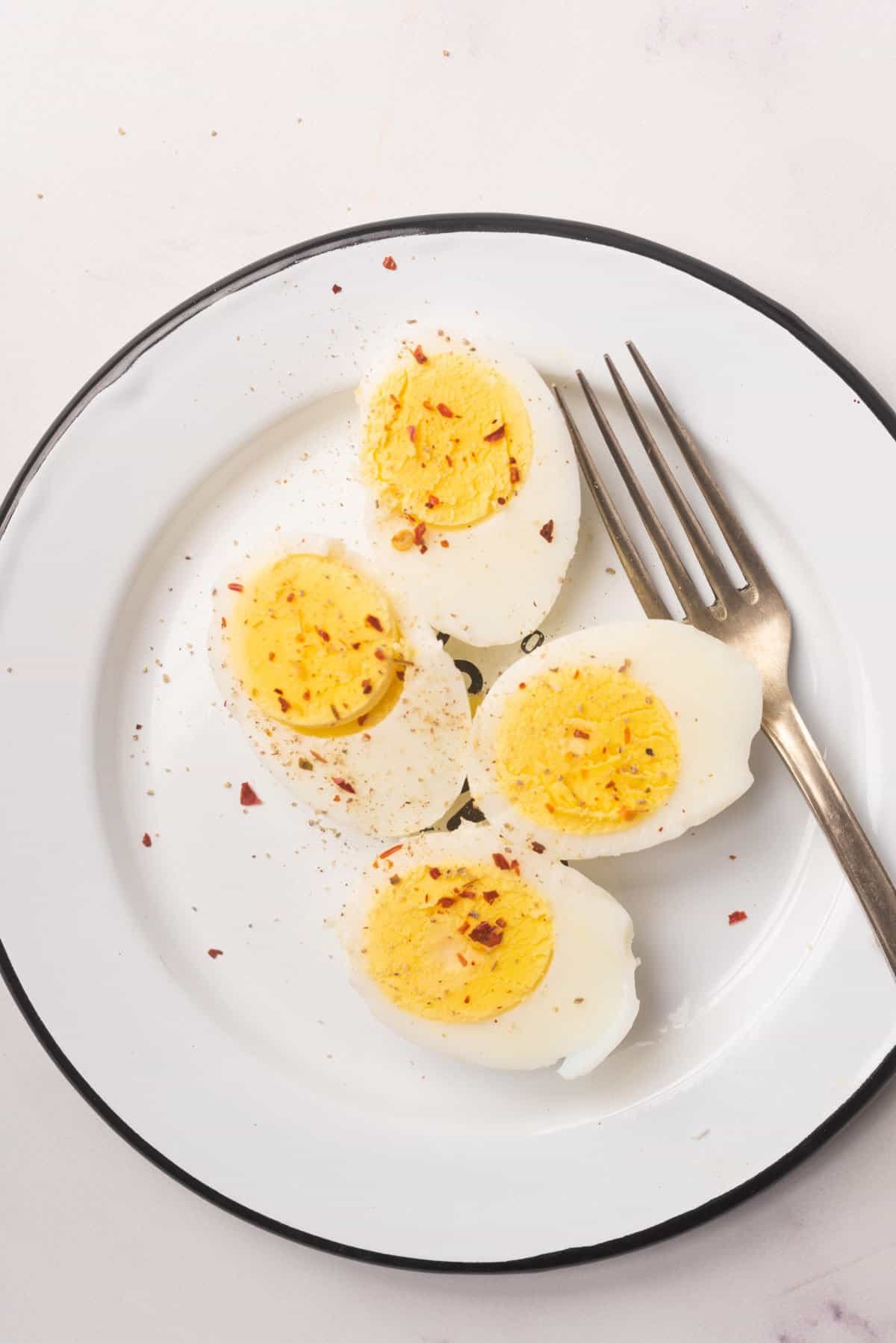 An image of two hard boiled eggs sliced in half, topped with seasonings, and being served on a plate with a fork beside them.