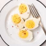 An overhead image of two hard boiled eggs sliced in half, topped with seasonings, and being served on a plate with a fork beside them.