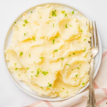 An image of microwave mashed potatoes in a bowl with fresh herbs on top and a fork resting on the right side of the bowl.