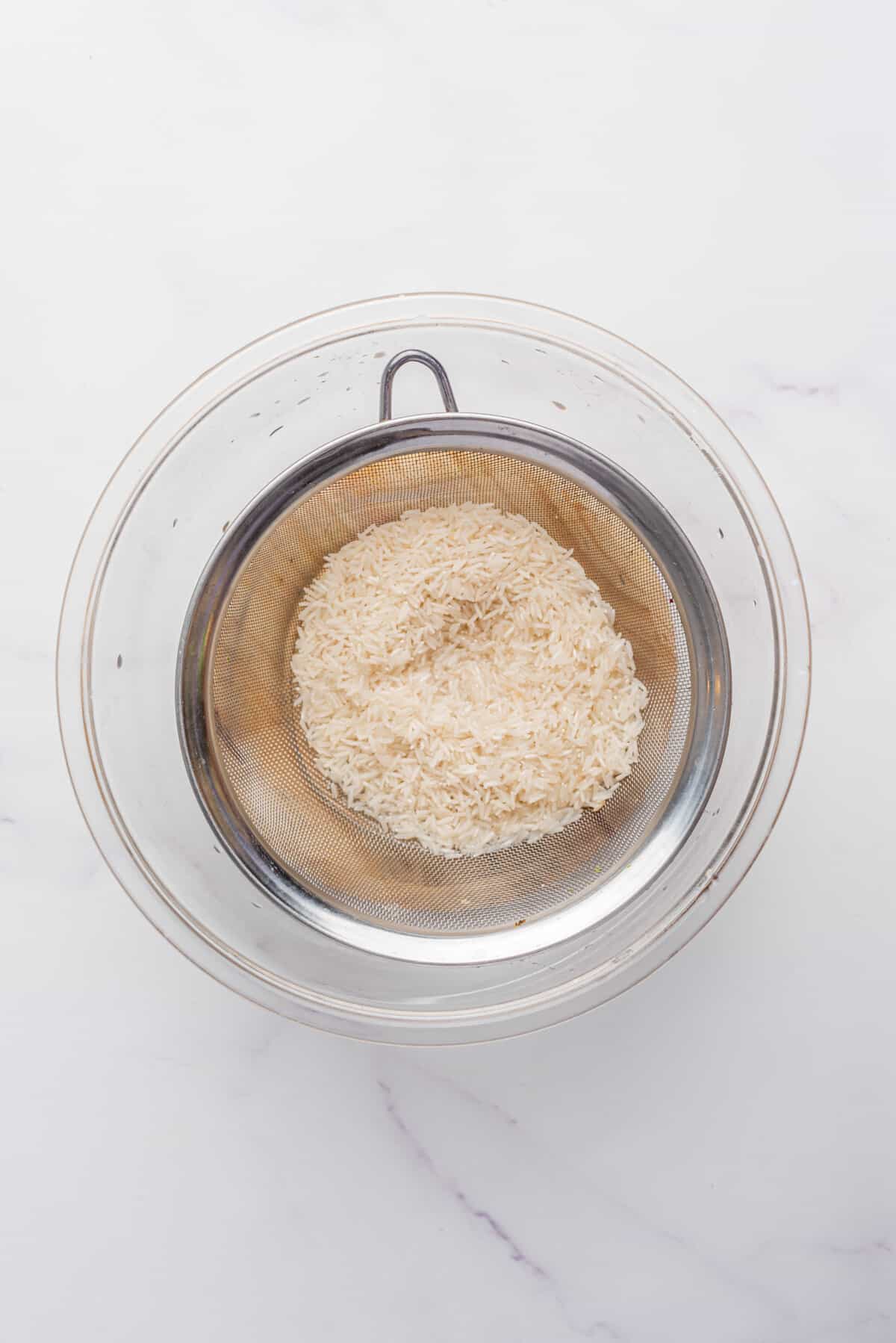 An overhead image of uncooked rice on a fine-mesh strainer inside of a bowl.