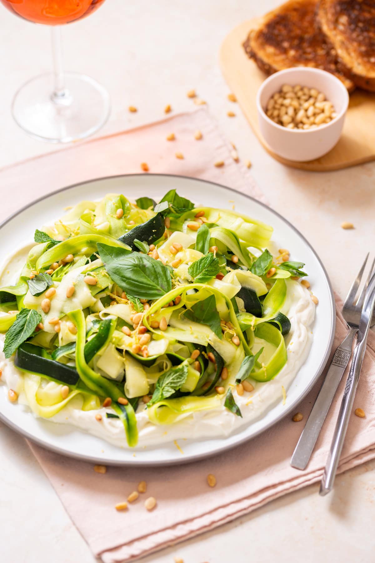 Zucchini ribbon salad over whipped lemon ricotta topped with basil, mint, toasted pine nuts, olive oil, and lemon zest on a white plate with toast and rosé nearby.