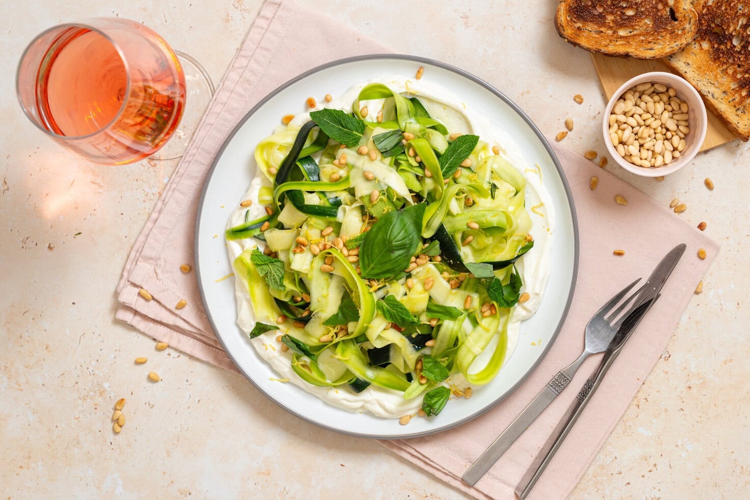 Zucchini ribbon salad over whipped lemon ricotta topped with basil, mint, toasted pine nuts, olive oil, and lemon zest on a white plate with toast and rosé nearby.