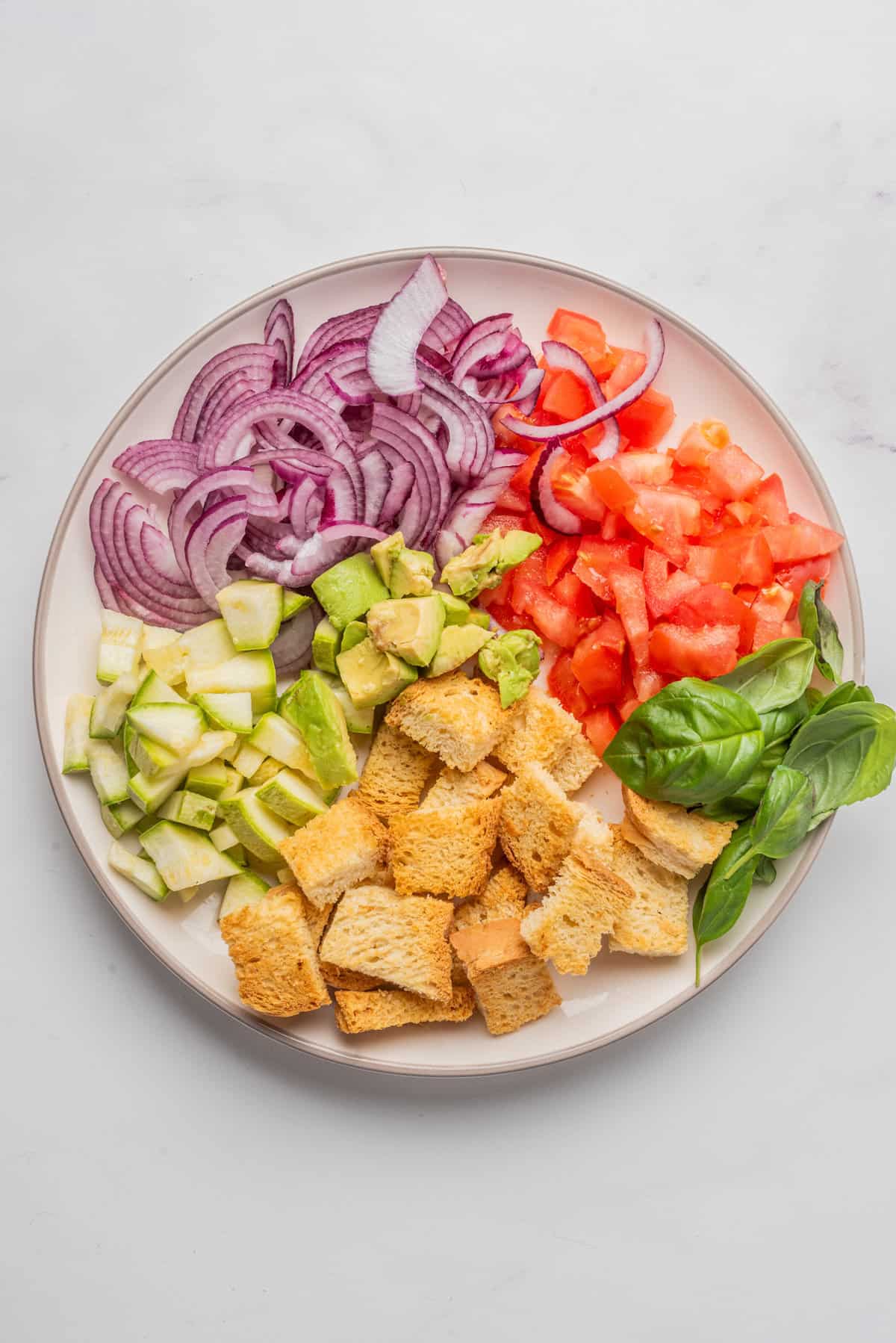 An overhead image of onions, tomatoes, avocadoes, crusty bread, zucchini, and fresh basil arranged side by side on a plate, before mixing together.