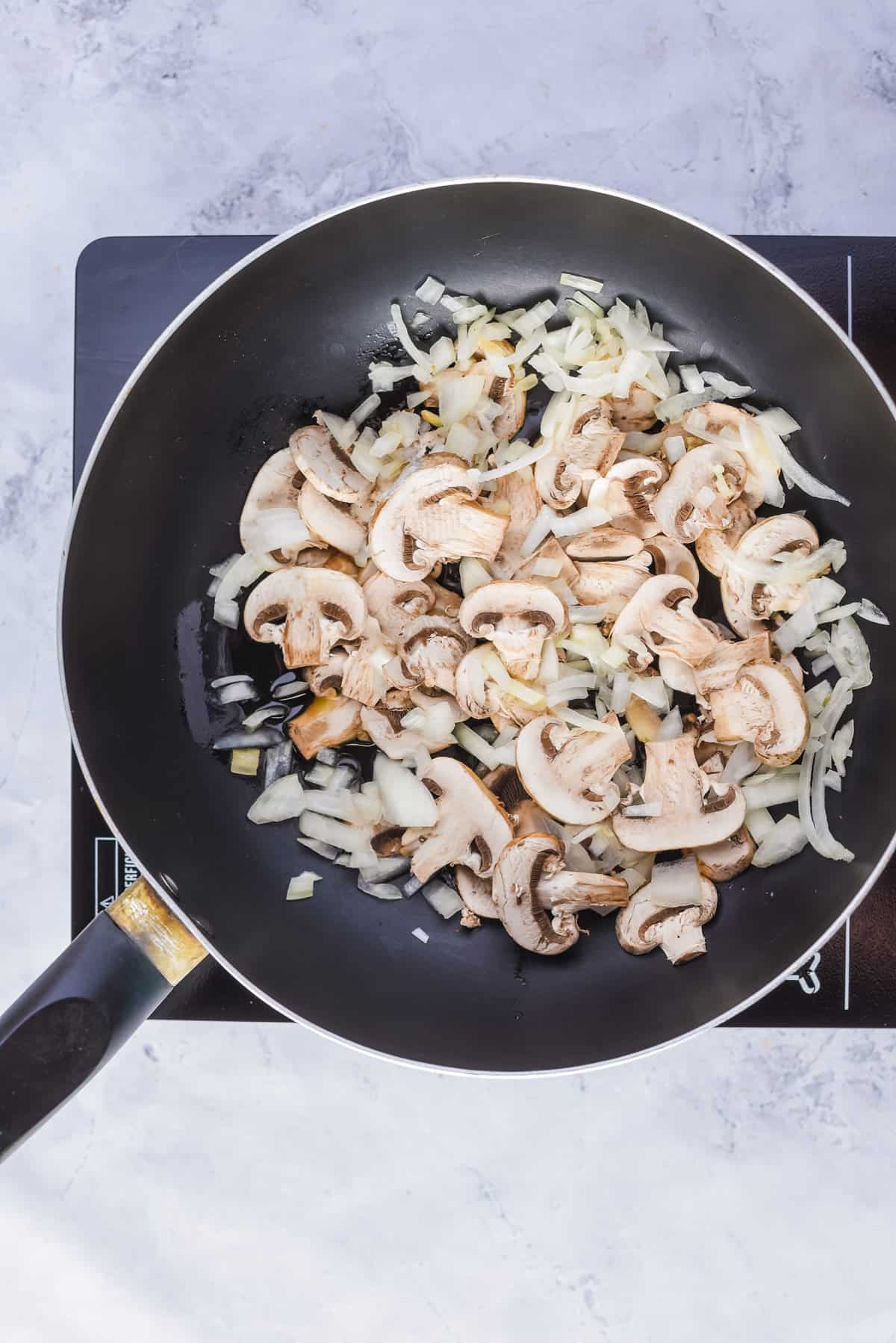 Overhead view of a skillet pan with oil, sliced onion, mushrooms, salt, and pepper.