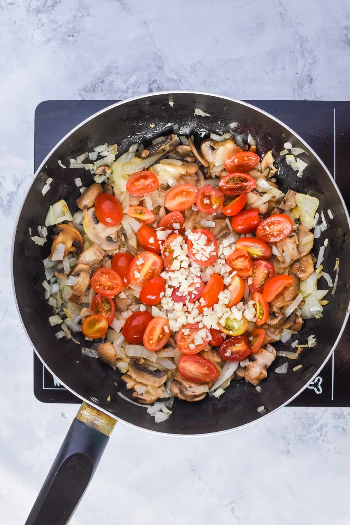 Overhead view of a skillet pan with oil, sliced onion, mushrooms, salt, and pepper added with cherry tomatoes and garlic.