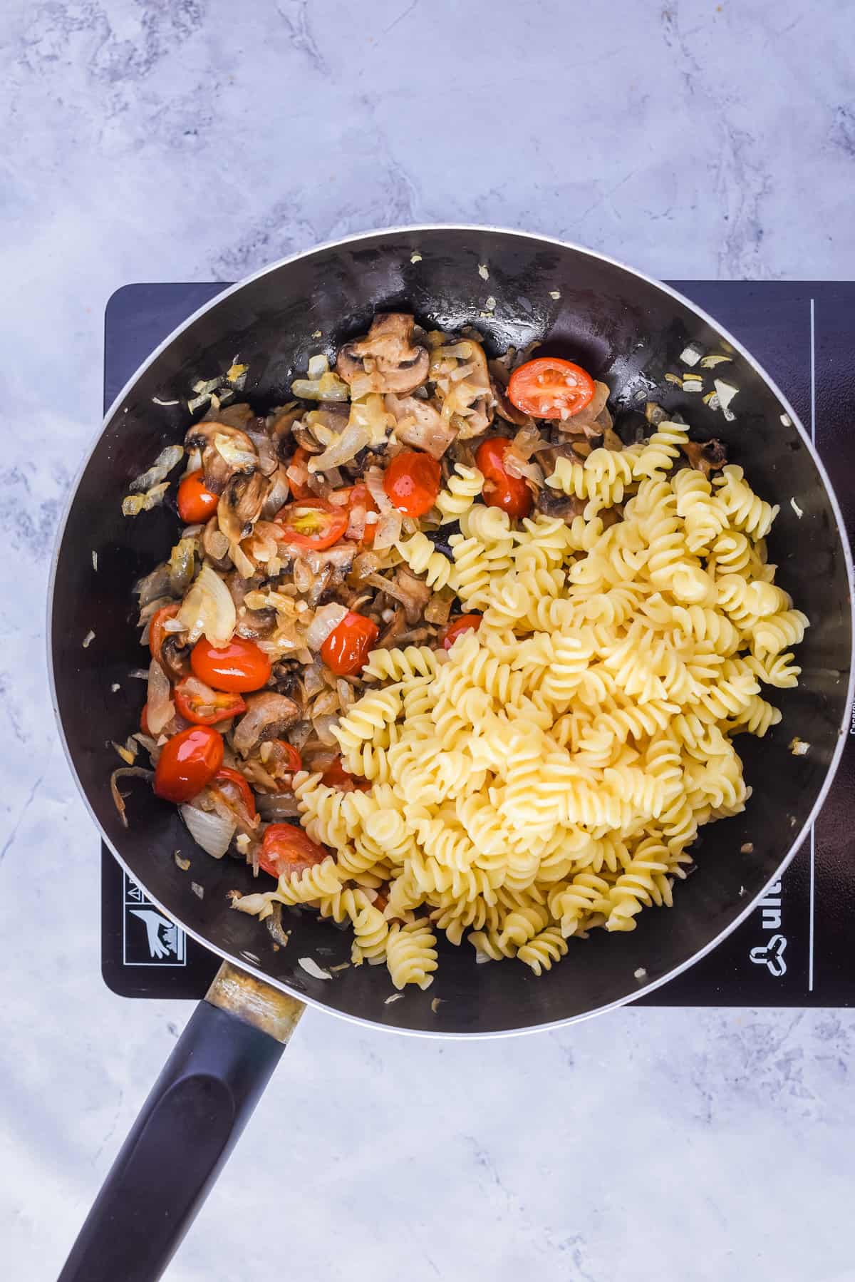 Overhead view of a skillet pan with oil, sliced onion, mushroom, salt, pepper, cherry tomatoes, and garlic added with pasta and butter.