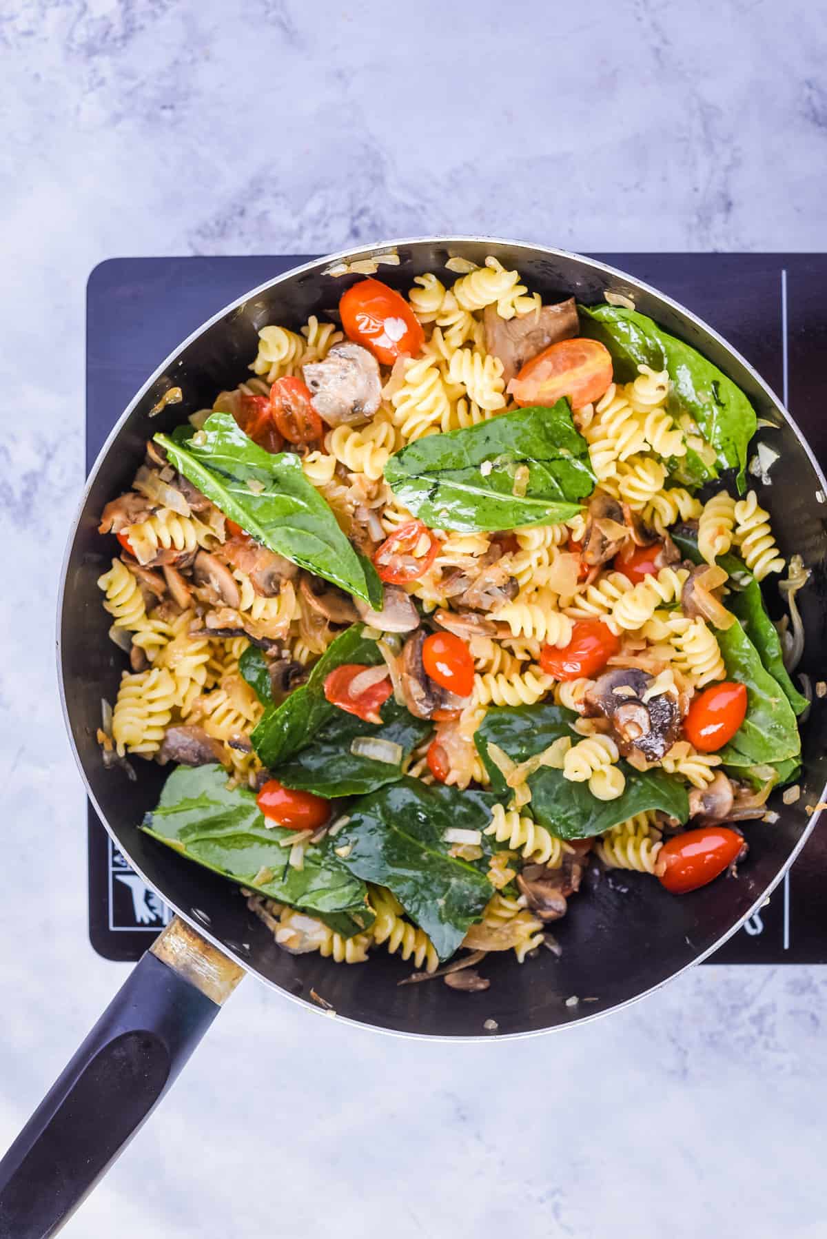Overhead view of a skillet pan with mixed oil, sliced onion, mushrooms, salt, pepper, cherry tomatoes, garlic, pasta, and fresh spinach.