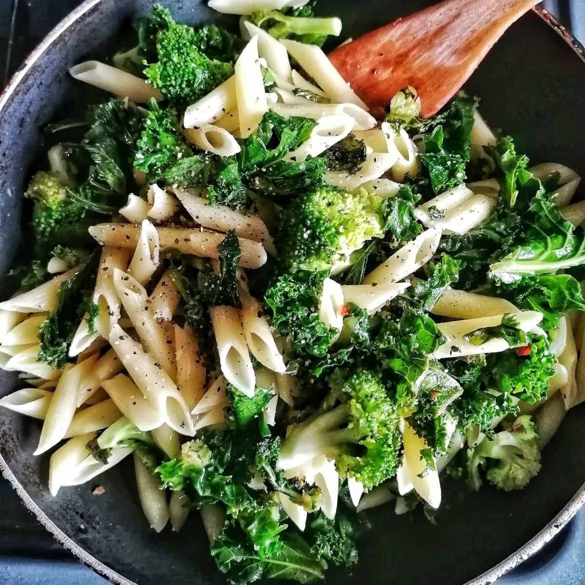 Overhead view of pasta salad with kale in a black bowl with a wooden serving spoon.