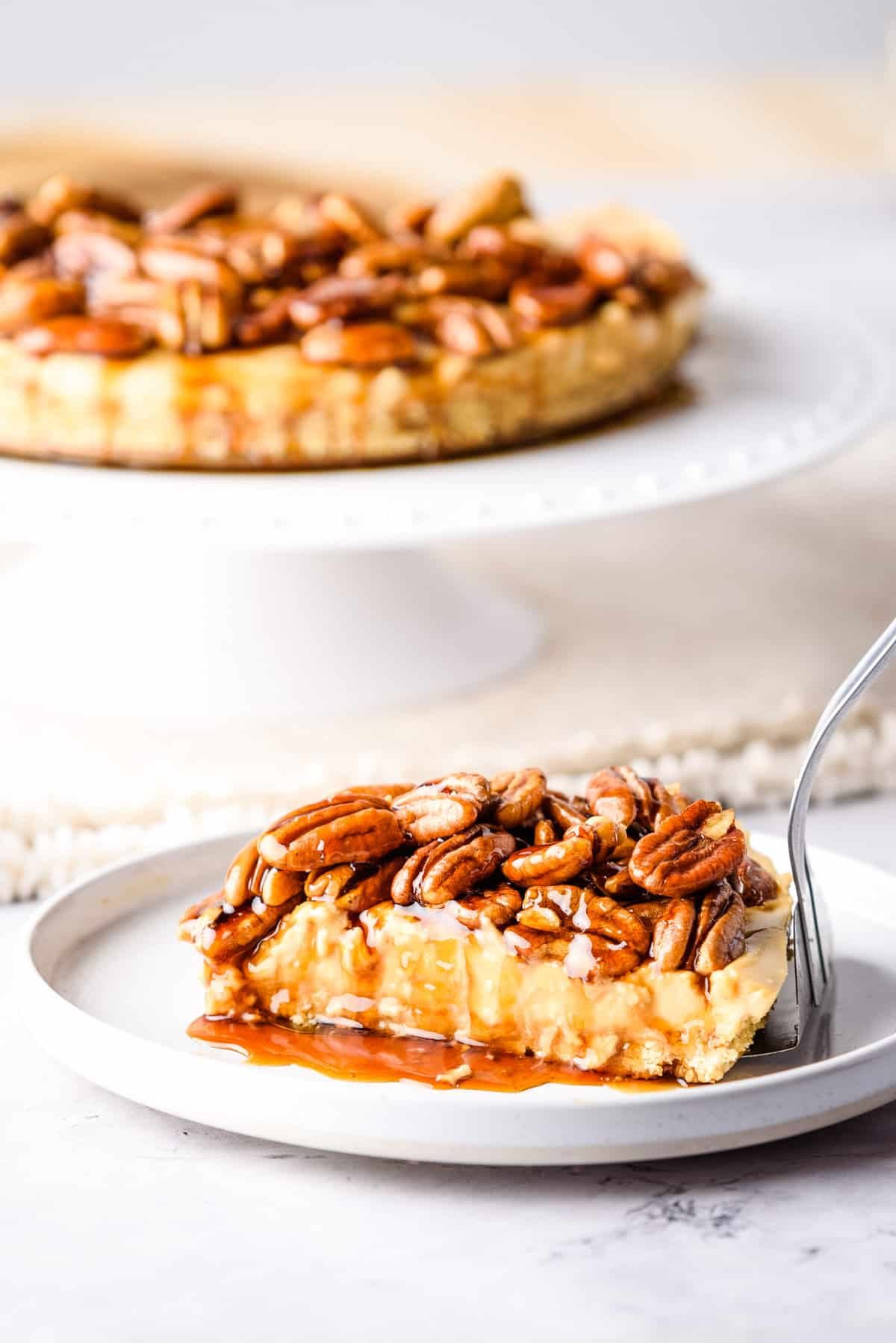 Close up of pecan pie cheesecake on a white plate, with the full cheesecake on a stand at the back.