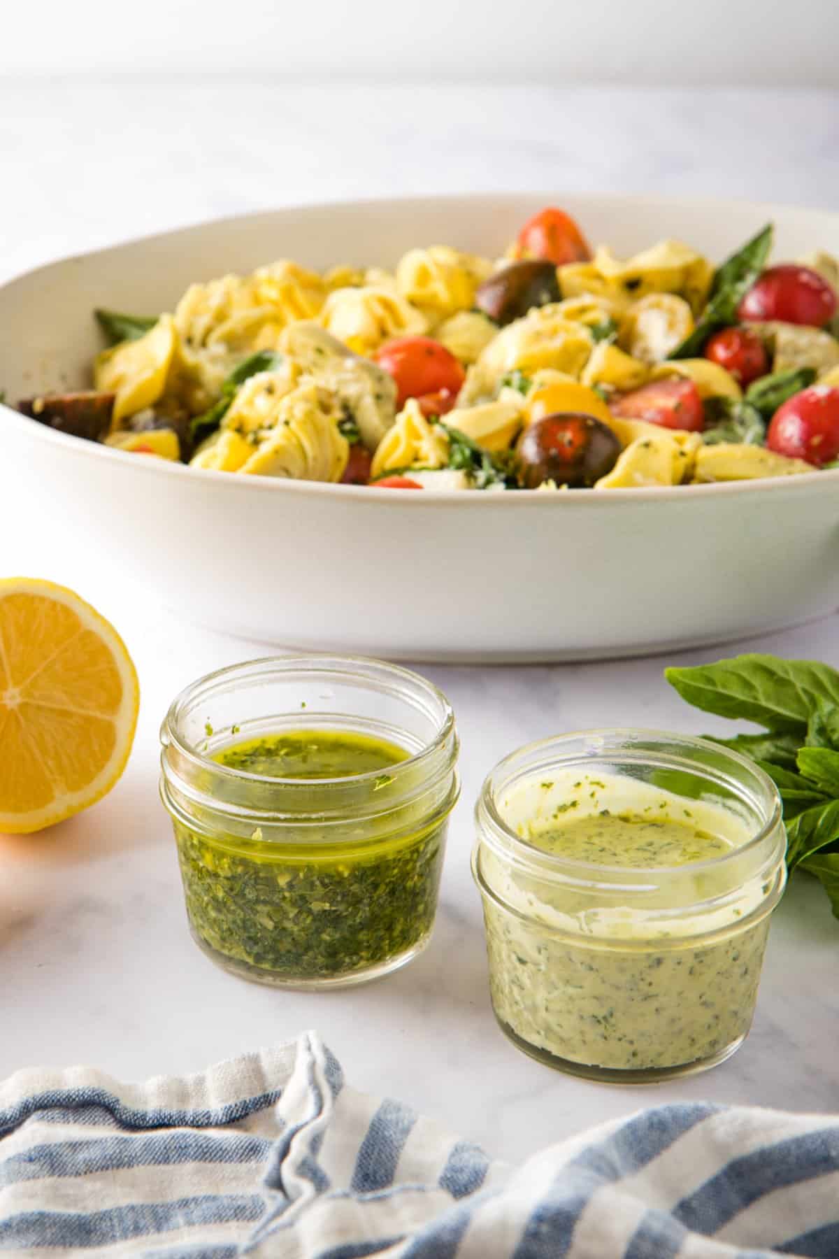 Picture of two types of pesto salad dressings in small jars, with a bowl of tortellini salad in the background.