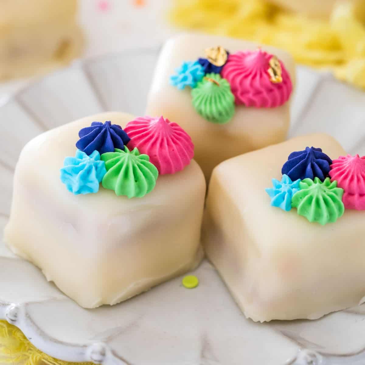 A close-up view of three petit fours placed on a white plate with colorful frosting on top.