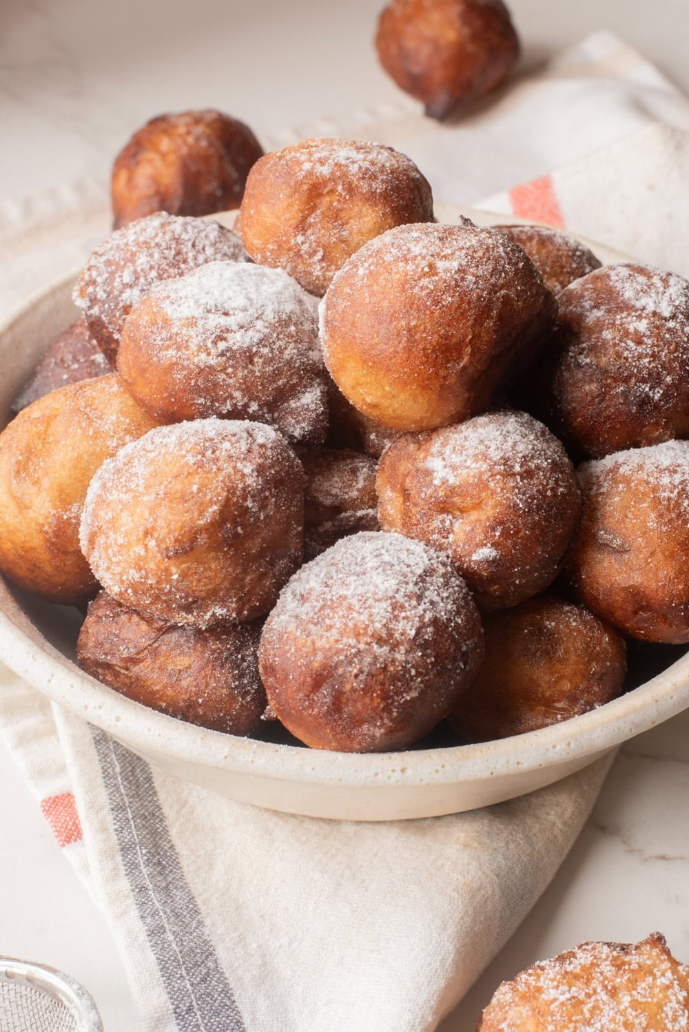 Bowl of golden-brown Nigerian puff puff dusted with powdered sugar on striped linen.