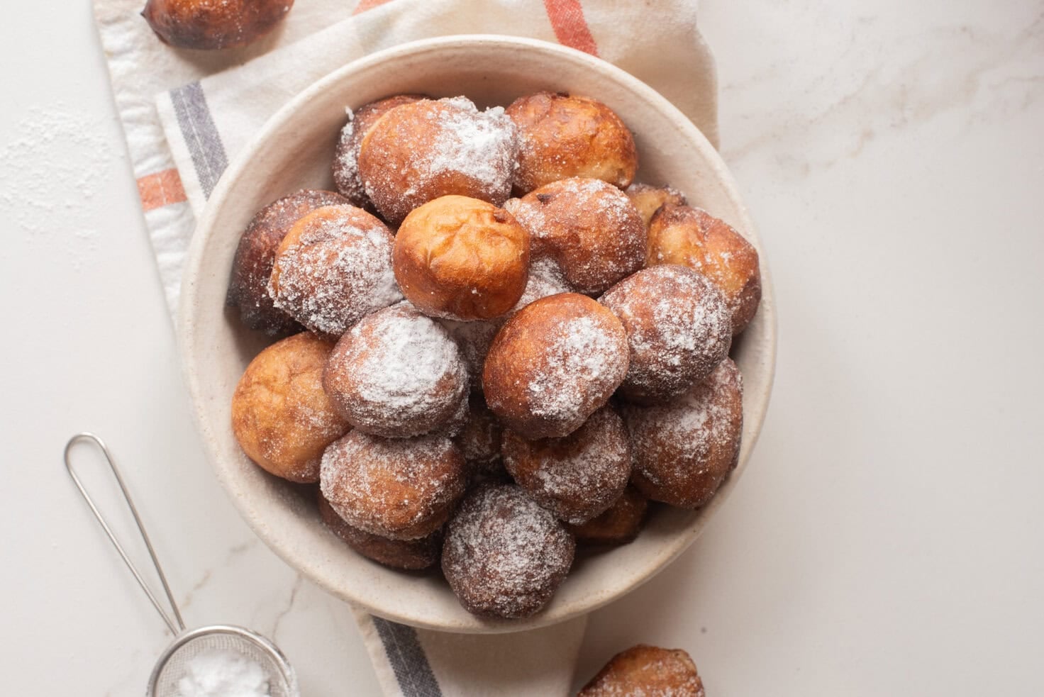 Overhead view of a bowl of Nigerian puff puff dusted with powdered sugar on a marble surface.