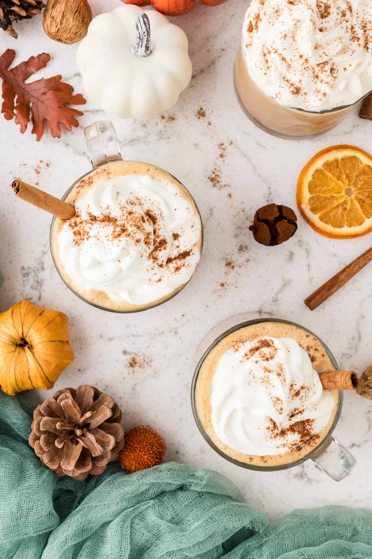 Two mugs of pumpkin spice latte with pumpkin and fall decor in the background.