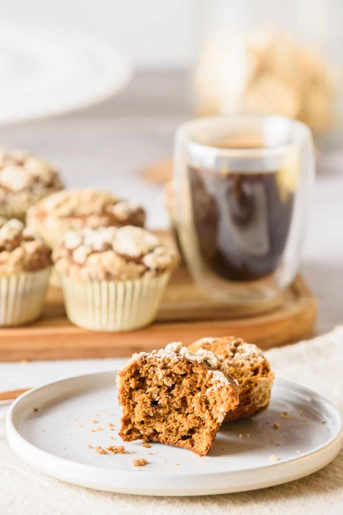 Pumpkin streusel muffin sliced in half, placed on a white plate, with a wooden platter of pumpkin muffins and coffee in the background.