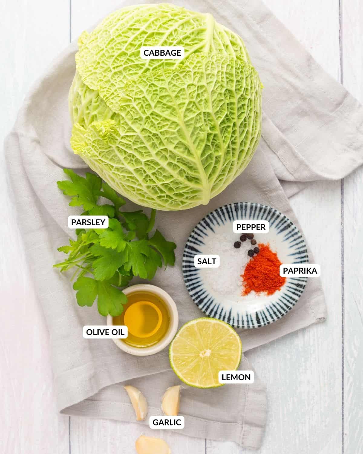 An overhead image of a head of cabbage, a slice of lemon, olive oil, garlic cloves, seasonings, and fresh parsley on a table with labels on each one.