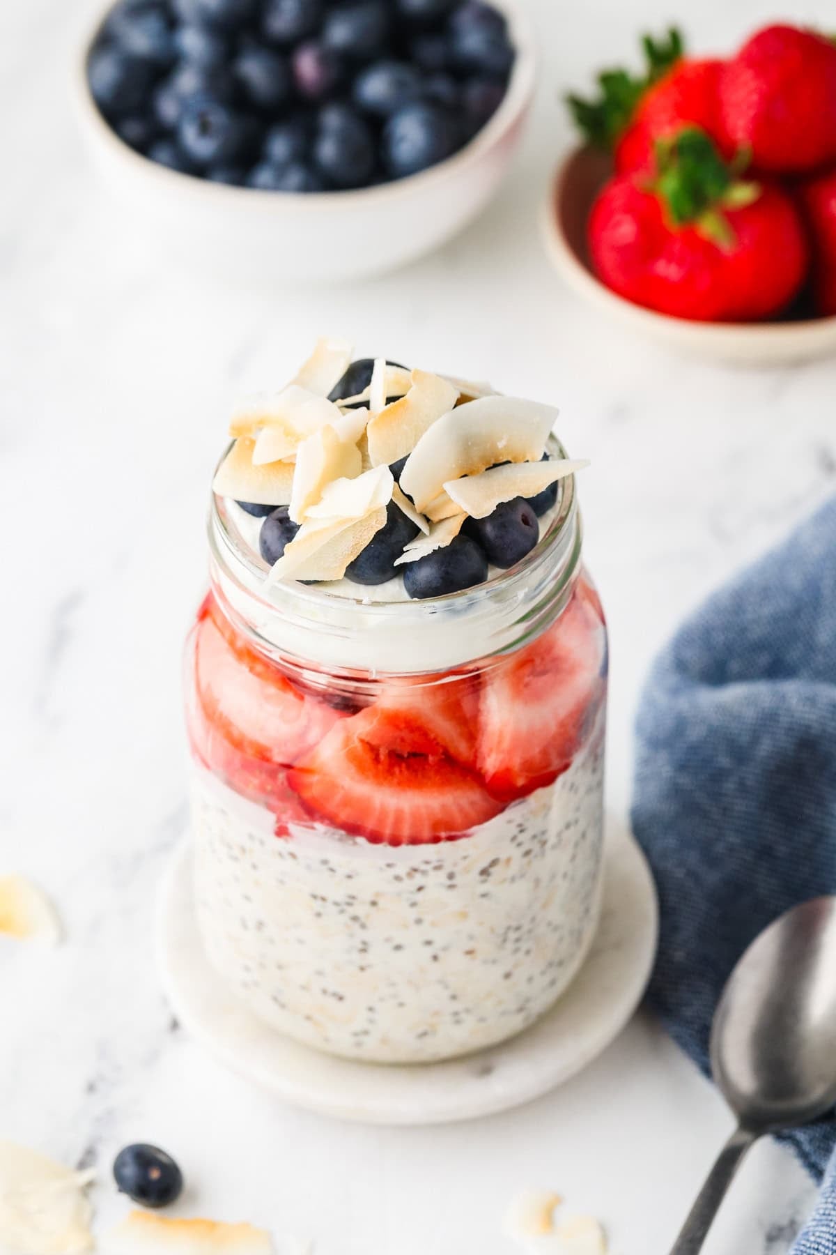 Red, White & Blue Overnight Oats in a mason jar topped with blueberries and shredded coconut, with fresh strawberries and blueberries in the background.