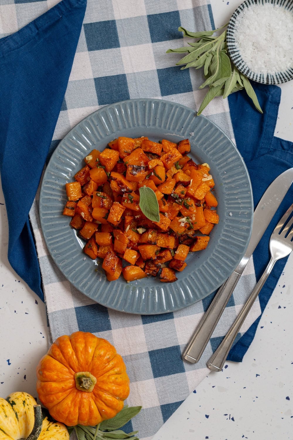Finished roasted butternut squash cubes with browned edges and fresh sage, served on a blue plate.