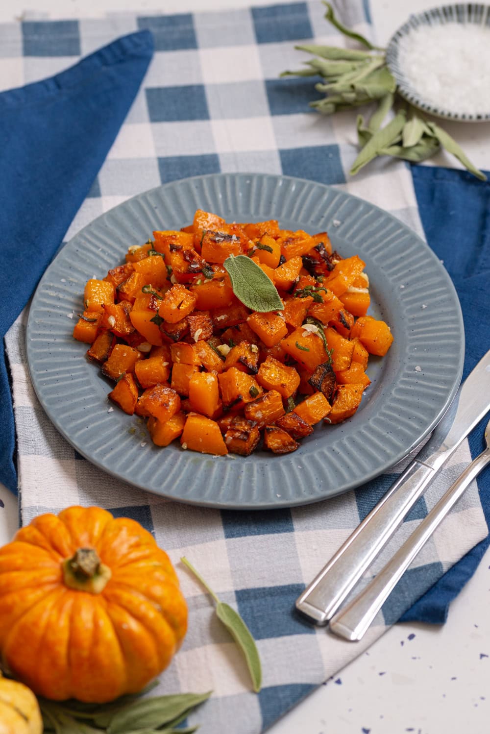 Roasted butternut squash cubes served on a blue plate with fresh sage and a small pumpkin.