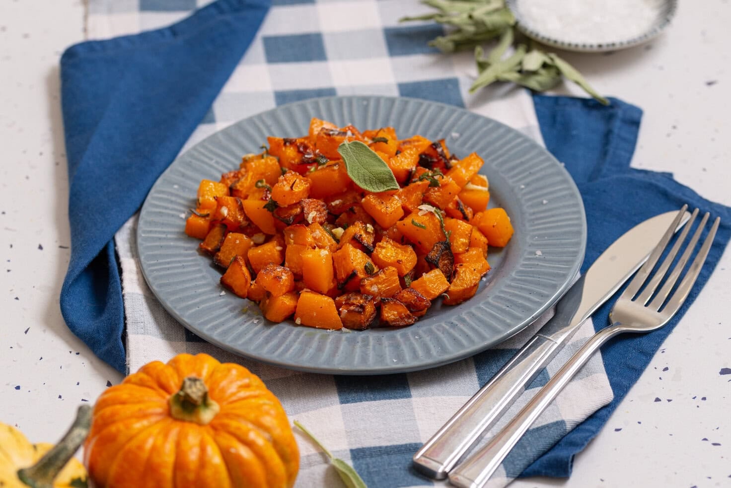 Roasted butternut squash cubes served on a blue plate with fresh sage and a small pumpkin.