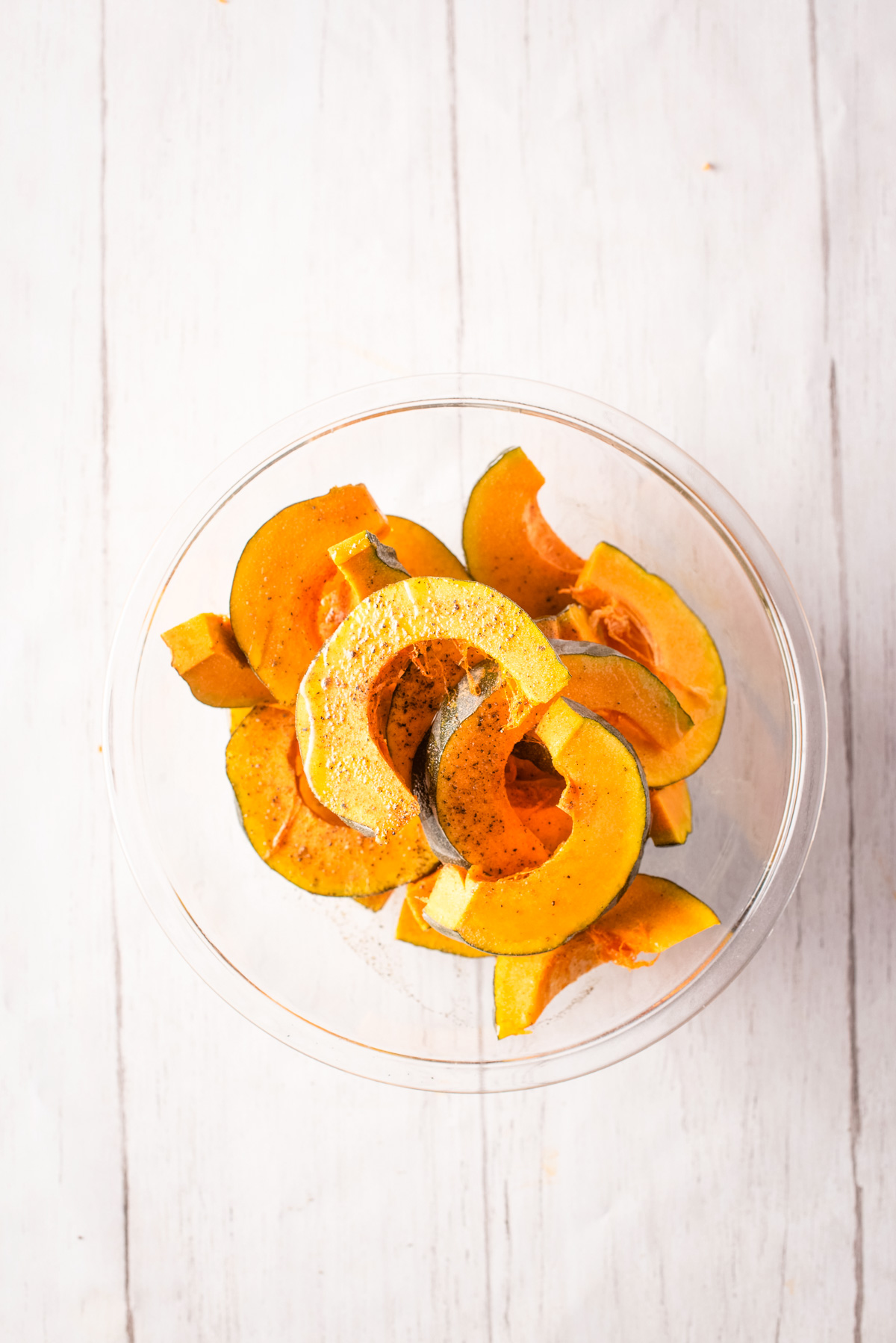 Overhead view of kabocha squash slices in a glass bowl tossed with salt and pepper.