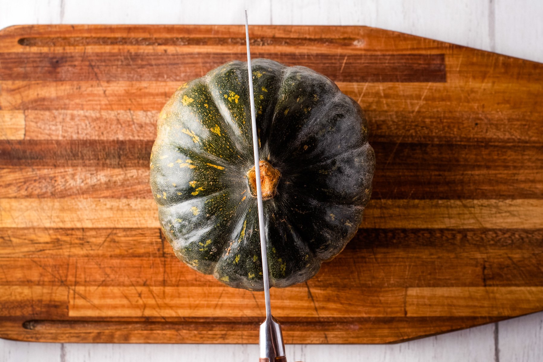 Overhead shot showing kabocha squash on a wooden cutting board, with knife positioned to cut it down the center.