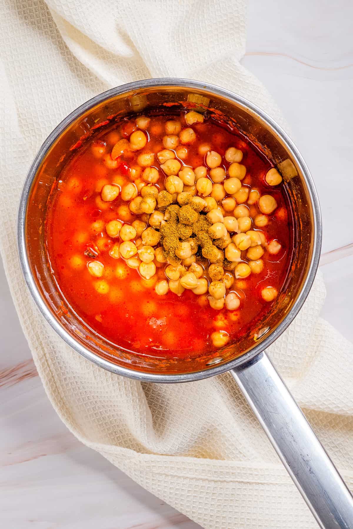Overhead view of a pan with stirred oil, cumin seeds, onions, garlic, ginger, chilies, turmeric, coriander, pureed tomatoes and salt added with chickpeas, water and garam masala.