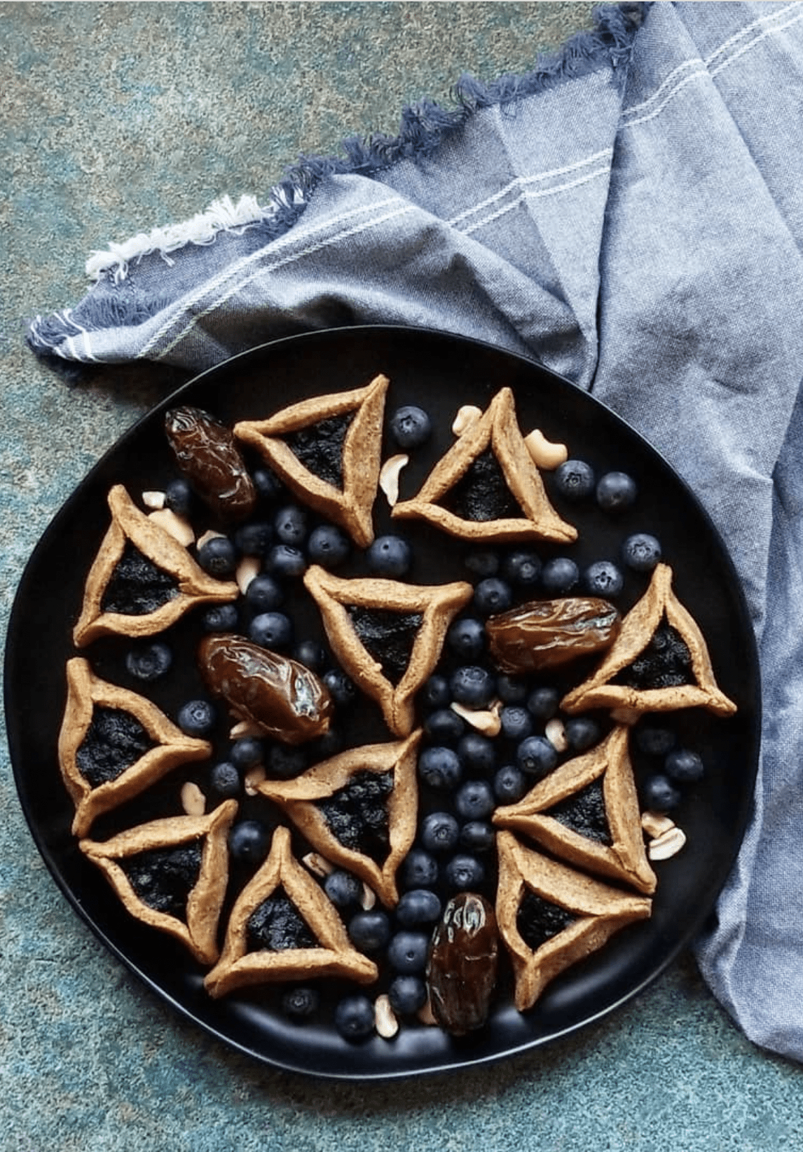 Overhead view of blueberry pie hamantashen cookies with large raisins, raw cashews, and blueberries on a black plate.