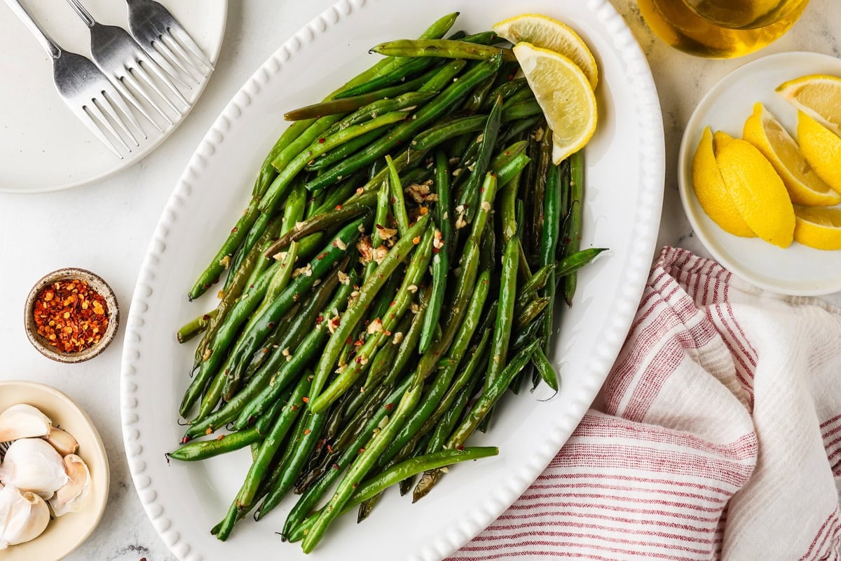 Platter of skillet green beans garnished with garlic, red pepper flakes, and fresh lemon wedges.