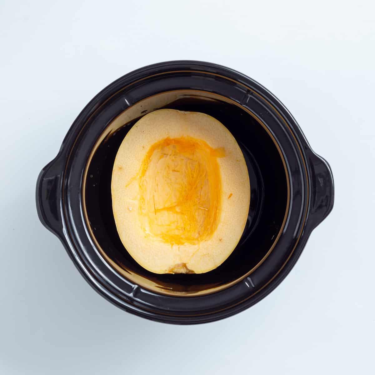 An overhead image of an uncooked spaghetti squash half in a slow cooker.
