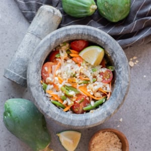 Vegan Thai green papaya salad served in a stone mortar with shredded papaya, carrots, green beans, tomatoes, peanuts, and lime.