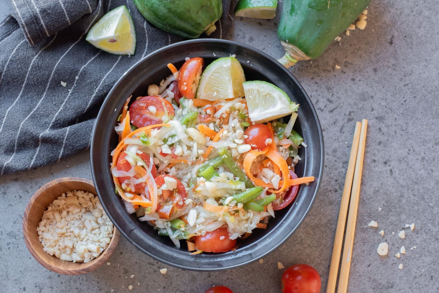 Thai green papaya salad with tomatoes, peanuts, and lime wedges served in a black bowl.