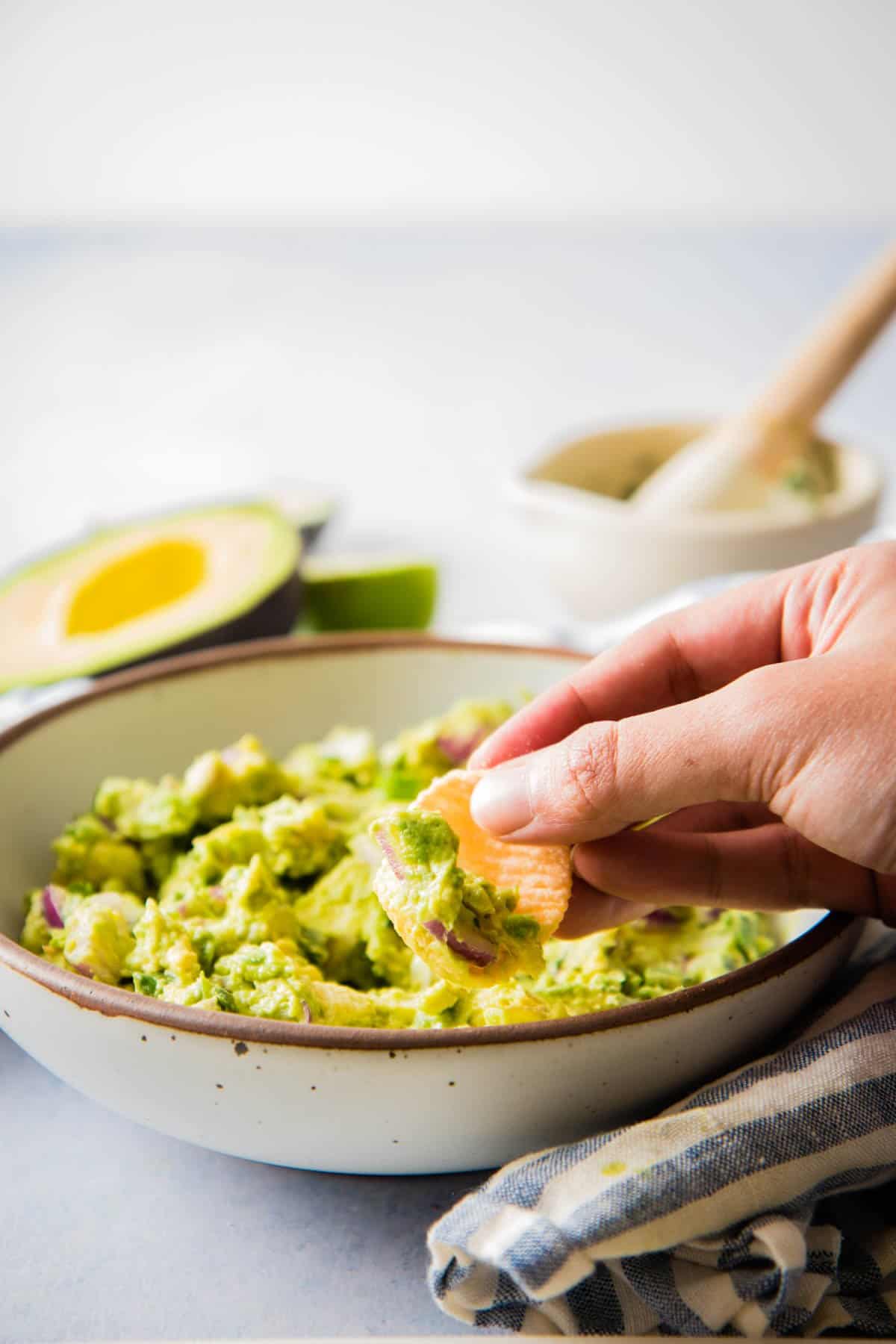 Close up of a a hand using a tortilla chip to scoop guacamole from a bowl 