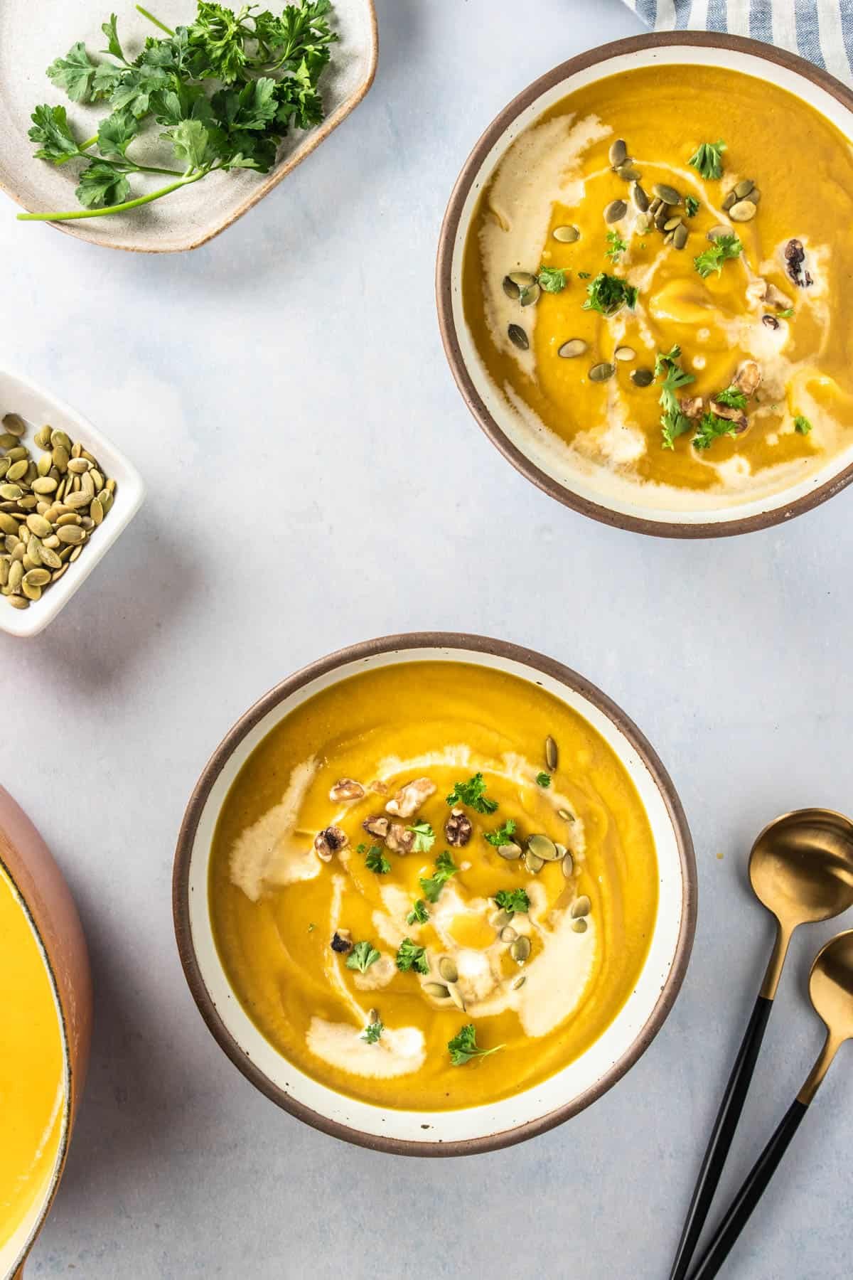 Two bowls of pumpkin soup garnishes with fresh herbs, coconut cream and toasted walnuts. 
