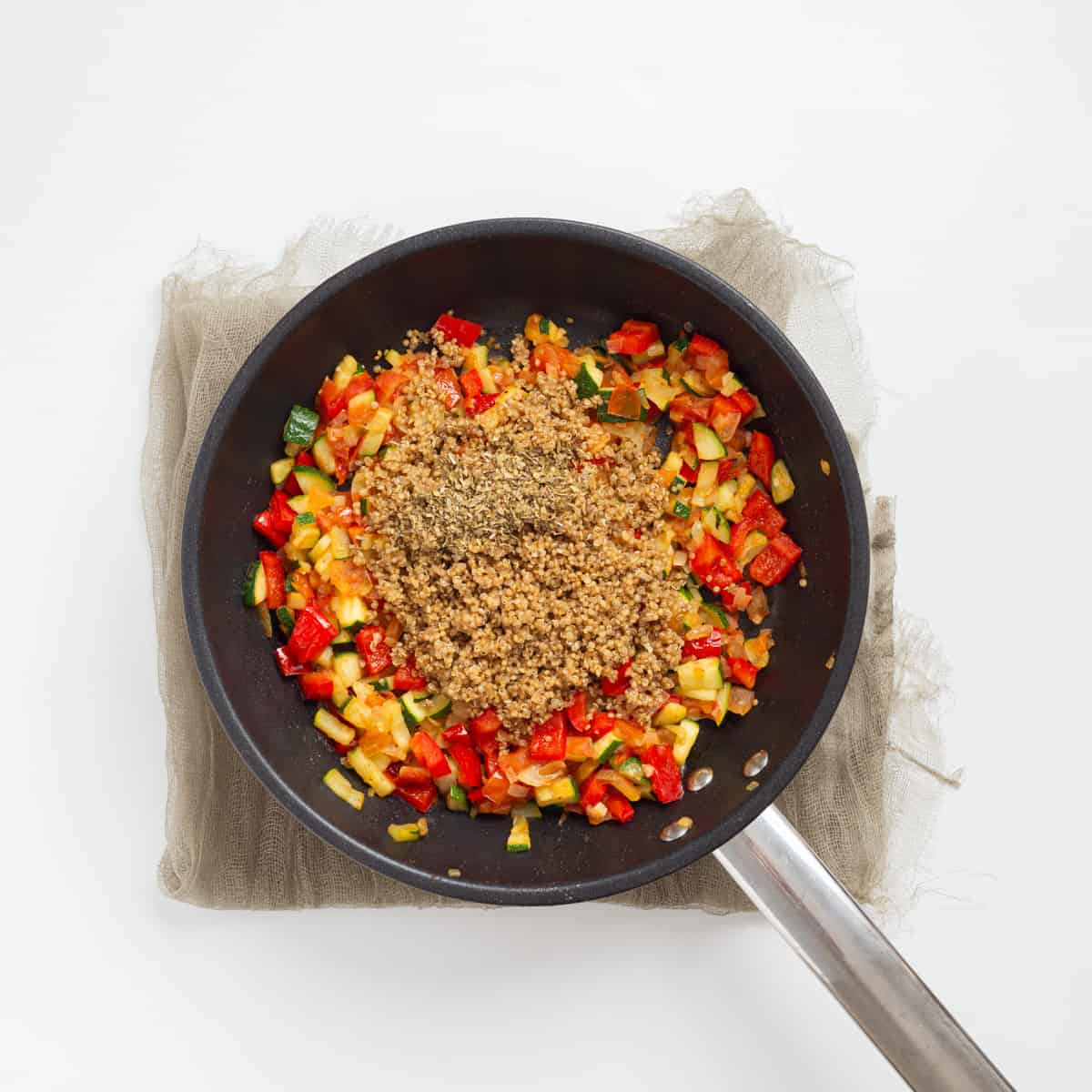 An overhead image of quinoa and dried italian seasoning being added to a skillet full of sauteed vegetables.