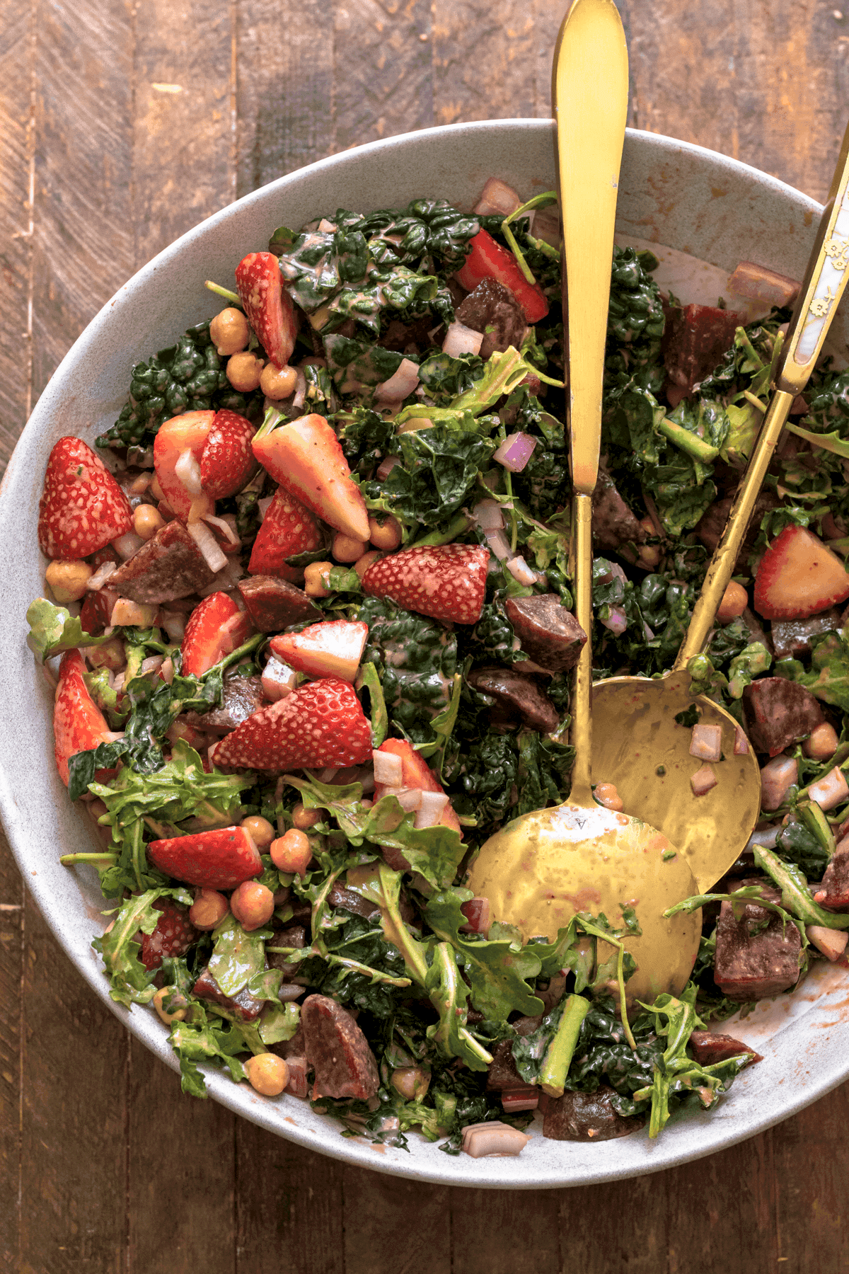 Overhead view of a strawberry kale salad in a white bowl with two gold serving spoons.