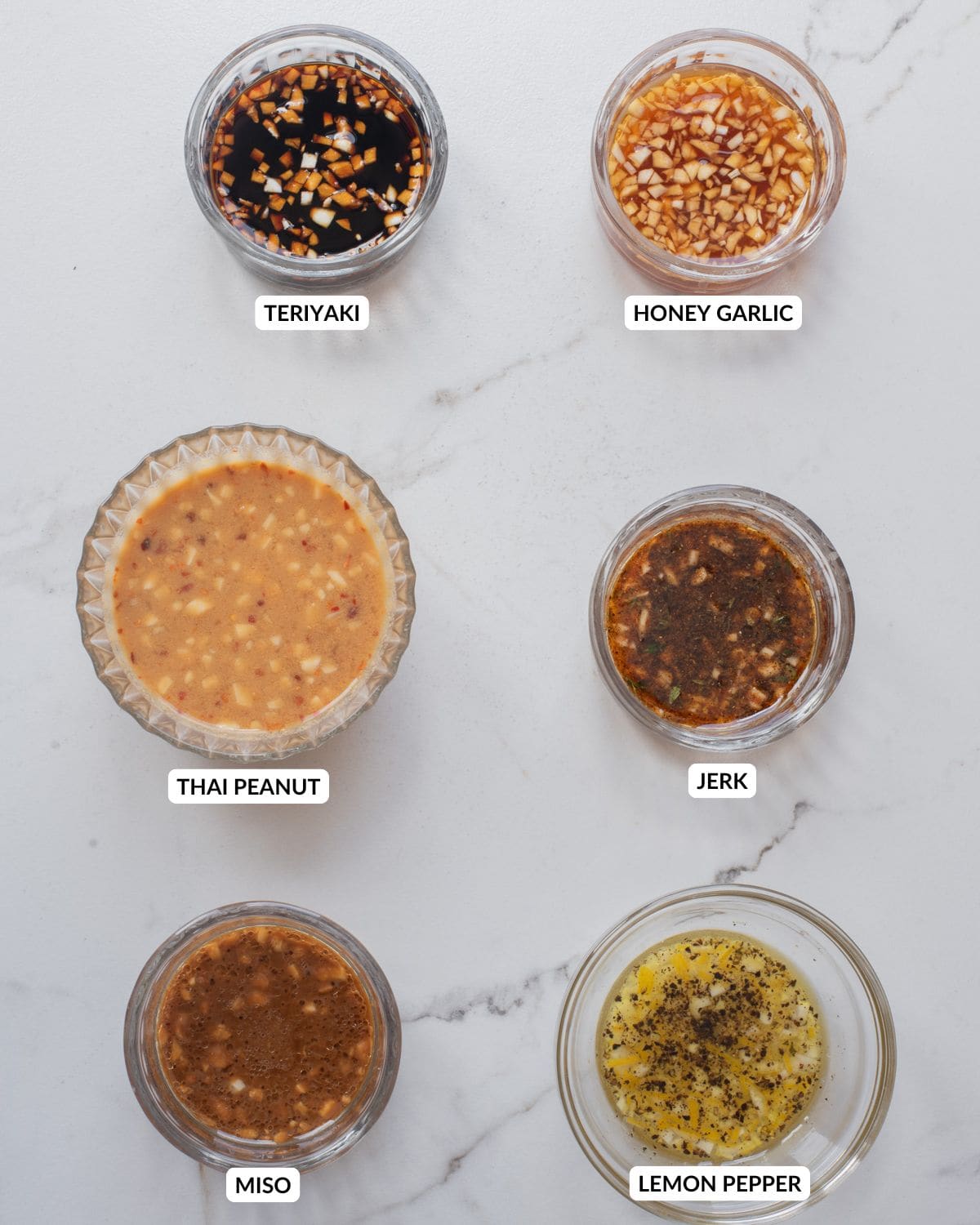 An overhead image of six different bowls with different marinades: teriyaki, honey garlic, jerk, miso, Thai peanut, and lemon pepper.
