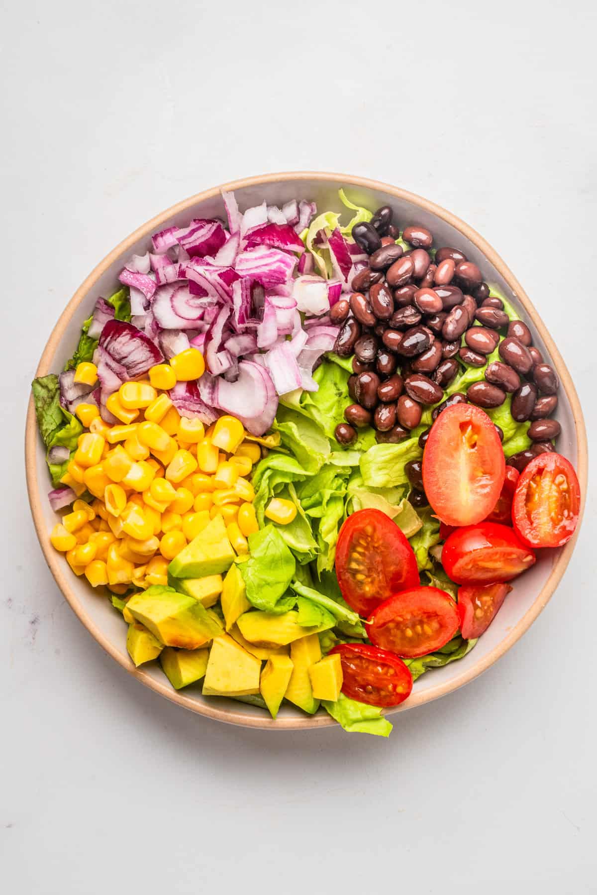 An overhead image of tomatoes, corn, onion, beans, sliced avocados, and onion arranged side by side in a bowl before mixing.
