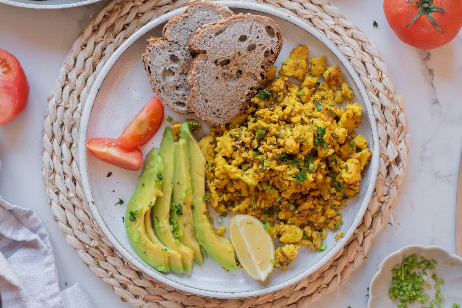 Plate of golden tofu scramble served with sliced avocado, toasted bread, and lemon wedges.